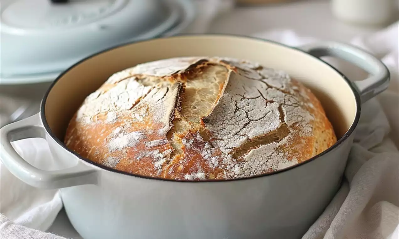 Golden brown round bread with a crackled flour-dusted crust in a pale blue Dutch oven on a white cloth, surrounded by wheat stems
