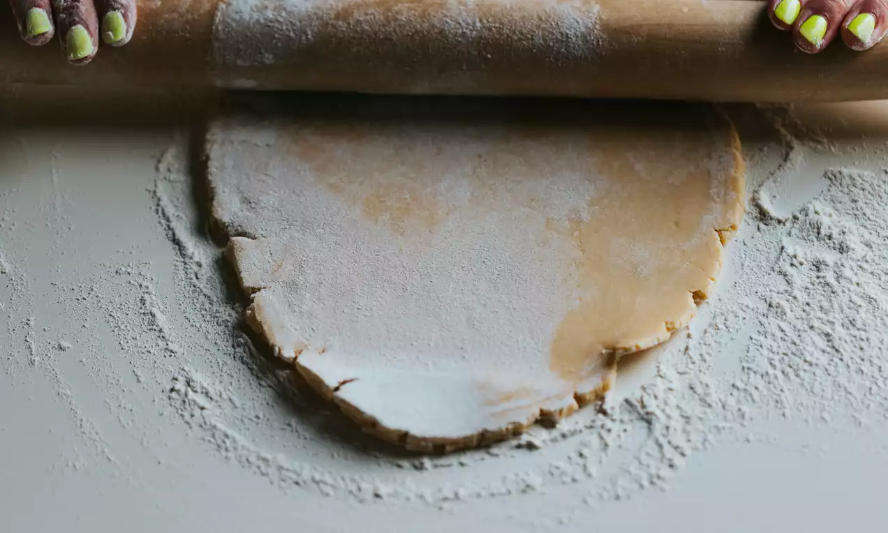 Pâte dorée et farineuse étalée en disque irrégulier sur une surface couverte de farine, roulée avec un rouleau en bois