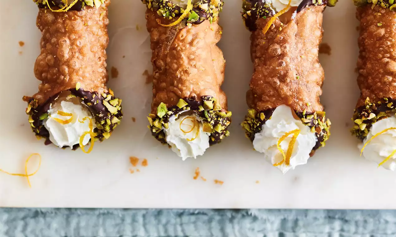 Cannoli dorés garnis de crème blanche, bords trempés dans du chocolat foncé et des pistaches, zeste de citron en décoration sur marbre blanc