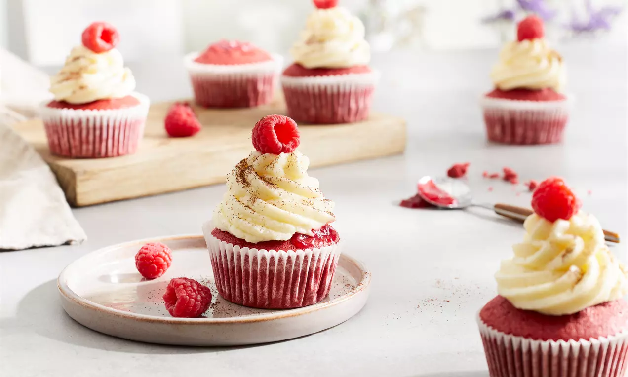 Petits gâteaux rouge velours surmontés de crème blanche tourbillonnée et décorés de framboises fraîches sur une table lumineuse