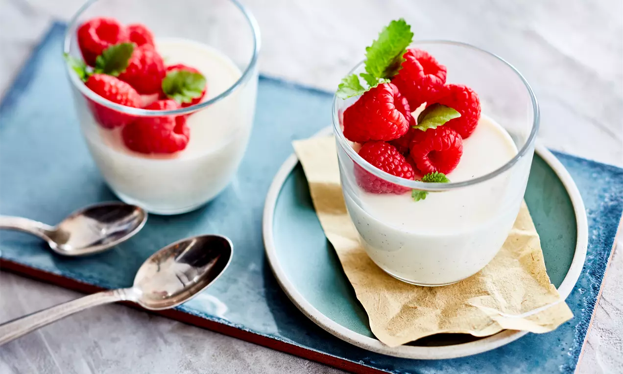Panna cotta à la vanille ivoire servie dans des verrines garnie de framboises rouges éclatantes et de feuilles de menthe verte