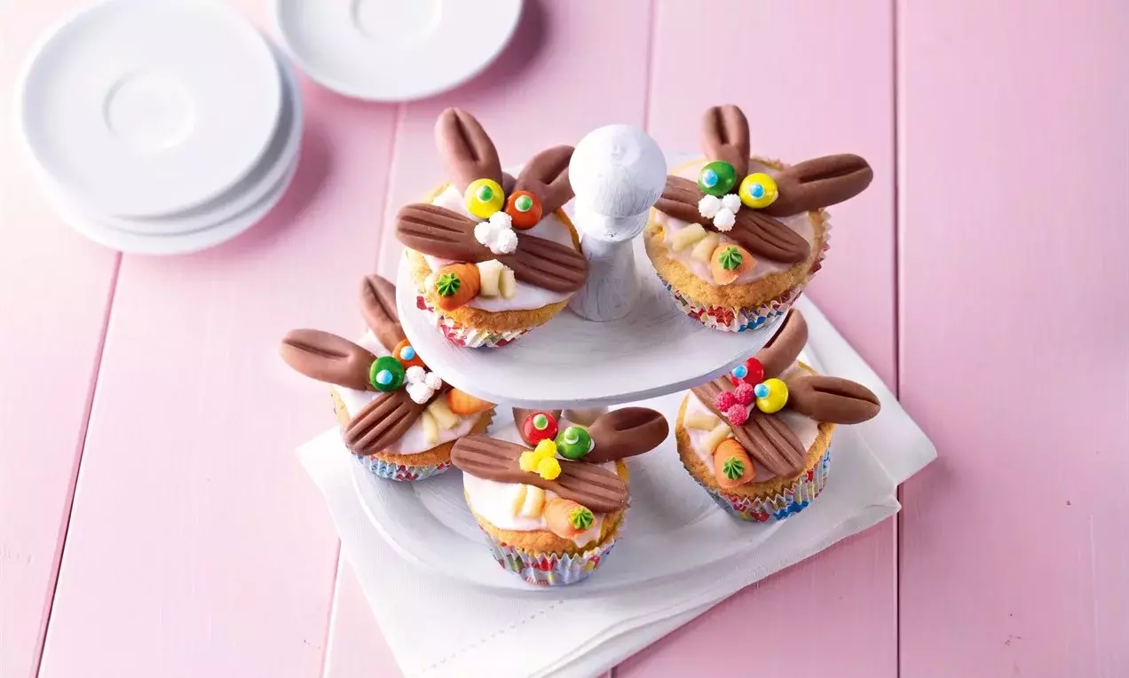 Petits cupcakes dorés décorés de glaçage coloré, oreilles en chocolat brun et détails en sucre sur un présentoir blanc à deux étages