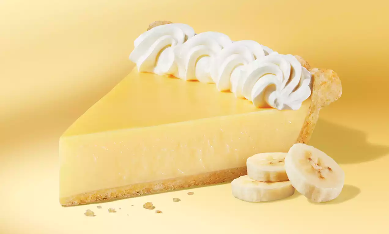 A slice of banana pie with glossy yellow filling on a pale crust, topped with white whipped cream swirls, surrounded by banana slices